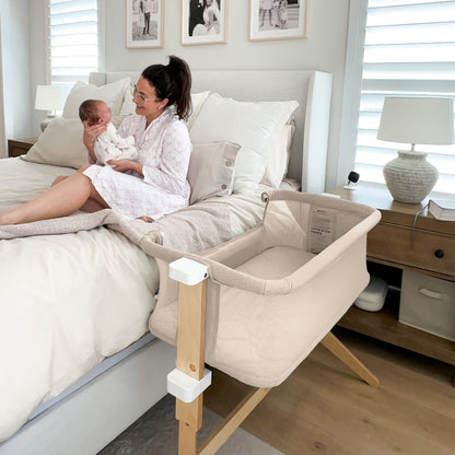 A woman in a robe sits on a bed holding a baby beside the Newton Baby Bassinet & Bedside Sleeper, which features breathable materials and wooden legs. The neutral room includes a nightstand with a lamp and framed photos for an inviting feel. 999-8400-SAN