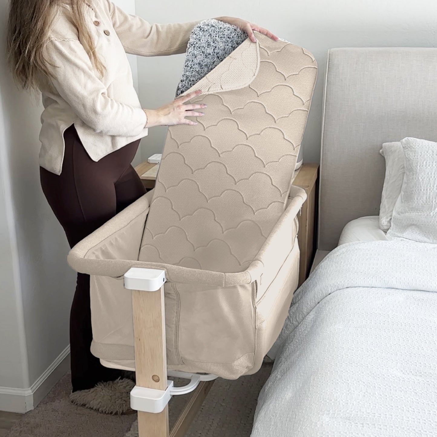 A woman puts a quilted beige mattress pad into the Newton Baby Bassinet & Bedside Sleeper, which is cream-colored, breathable, and positioned next to a bed in a softly lit bedroom.