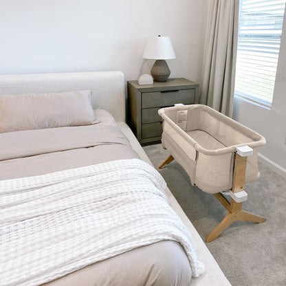 A cozy bedroom features a beige bed, white textured blanket, bedside table with lamp, and the Newton Baby Bassinet & Bedside Sleeper with wooden legs next to the bed. Sunlight streams in through window blinds.