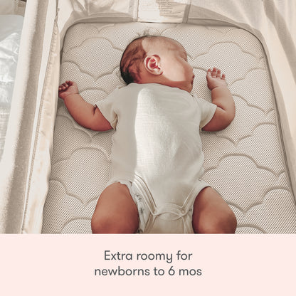 A baby in a white onesie sleeps on their back in the Newton Baby Bassinet & Bedside Sleeper. Text below: Extra roomy for newborns to 6 months.