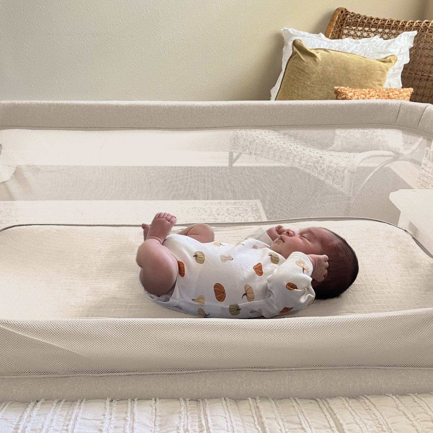 A newborn sleeps peacefully in a beige bedside sleeper, cozy on Newton Baby Bassinet Extra Sheets made of soft cotton muslin, with pillows and a woven chair in the background.