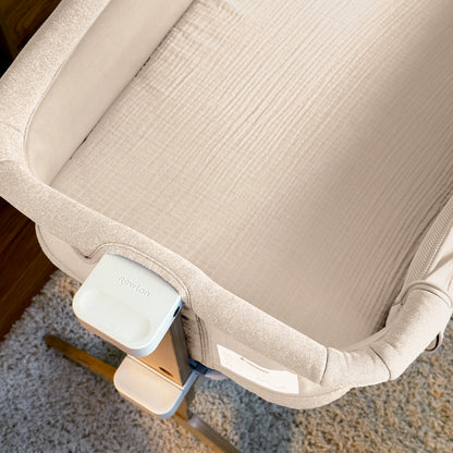 A close-up of Newton Baby Bassinet Extra Sheets in beige on a textured mattress, attached to a wooden support and set on light carpet. The Newton Baby brand is visible on the white latch.