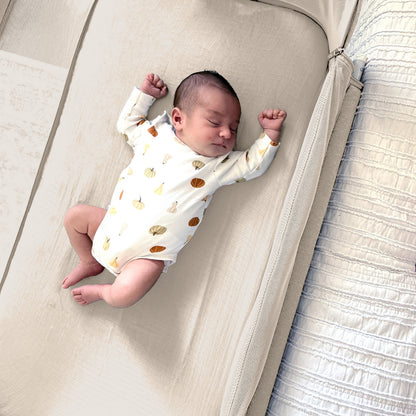 A newborn sleeps on a beige crib mattress, wearing a white onesie with colorful fruit prints. Soft Newton Baby Bassinet Extra Sheets made from cotton muslin surround the bedside sleeper for added comfort.