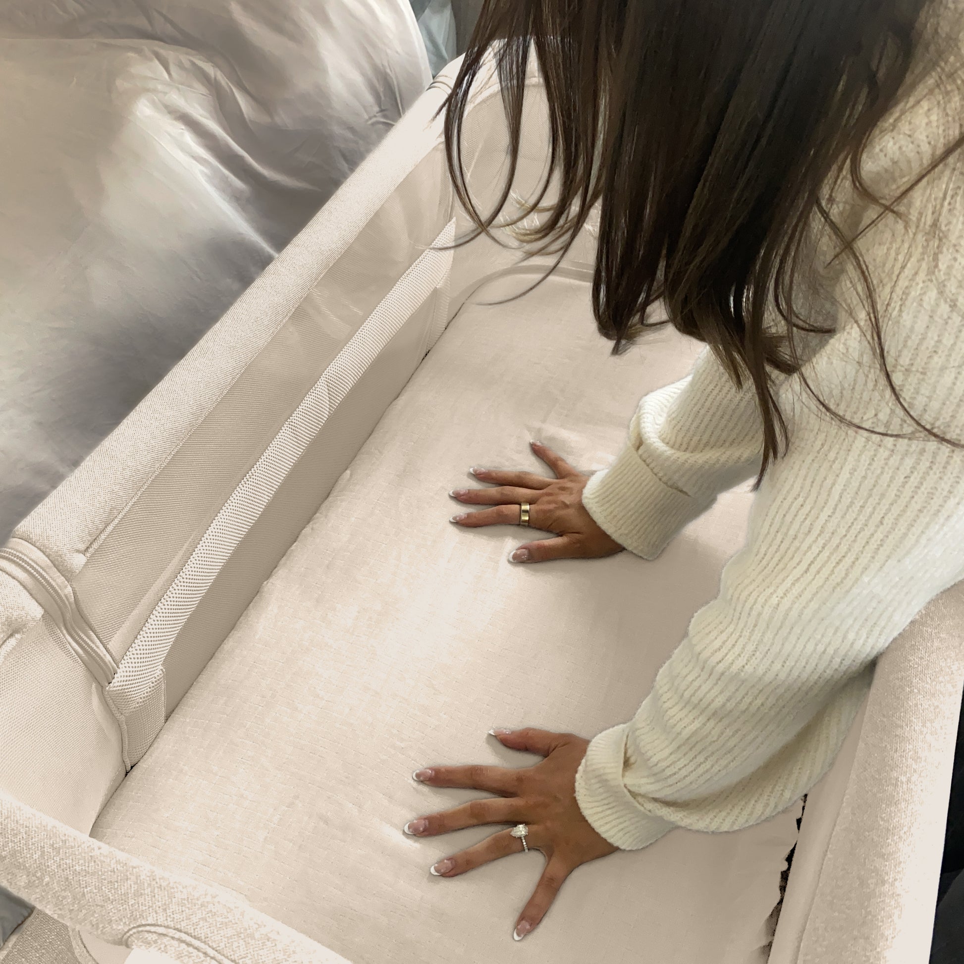 A person with long brown hair and a cream sweater presses down on a beige bassinet mattress, testing its firmness, while Newton Baby’s Bassinet Extra Sheets provide extra comfort with their soft cotton muslin fabric.