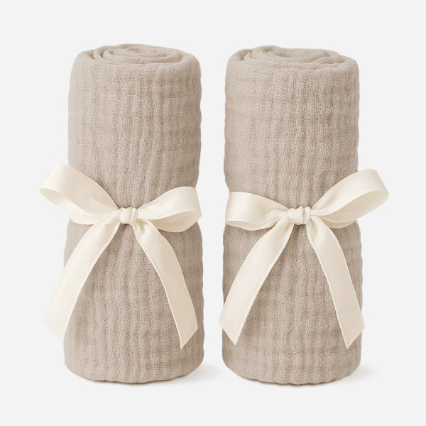 Two beige, waffle-textured cotton muslin Bassinet Extra Sheets from Newton Baby are rolled up and tied with cream satin ribbons, displayed upright against a plain white background.