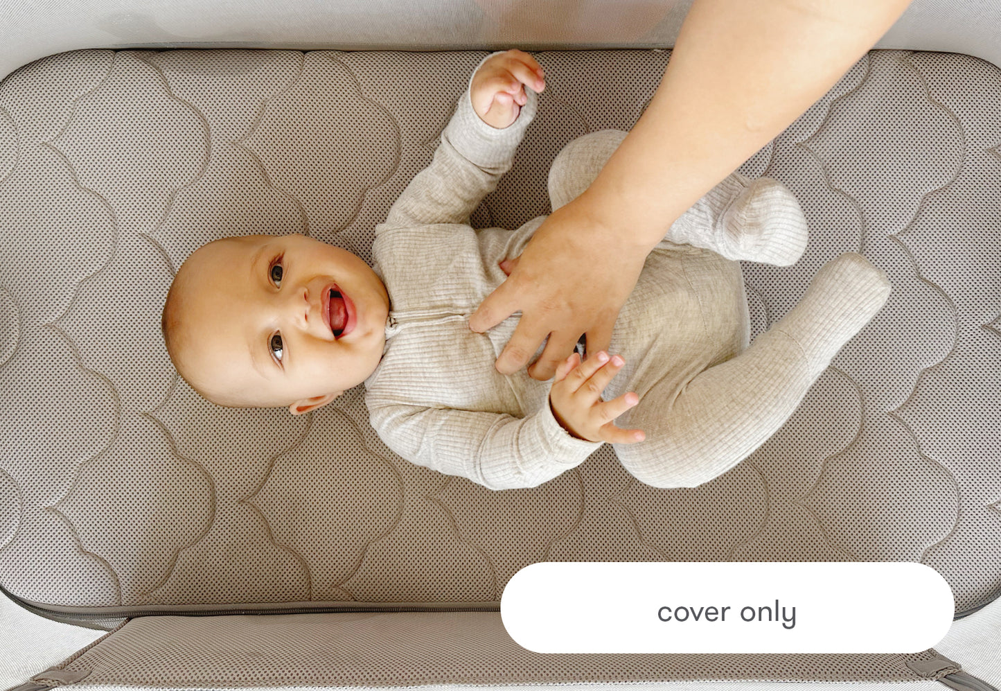 A joyful baby lying on a textured bassinet cover labeled "cover only", smiling while playing with an adult's hands.