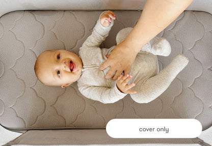 A joyful baby lying on a textured bassinet cover labeled "cover only", smiling while playing with an adult's hands.