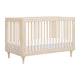 The Babyletto Lolly Convertible Crib features a minimalist design with vertical slats on all sides, four slightly tapered legs, and is crafted from natural New Zealand pine wood with a white mattress inside. skip