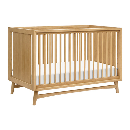 The Babyletto Peggy Convertible Crib features vertical slats, a white mattress, and angled legs. With a natural wood finish and minimalist mid-century style, it's ideal for any modern nursery. skip