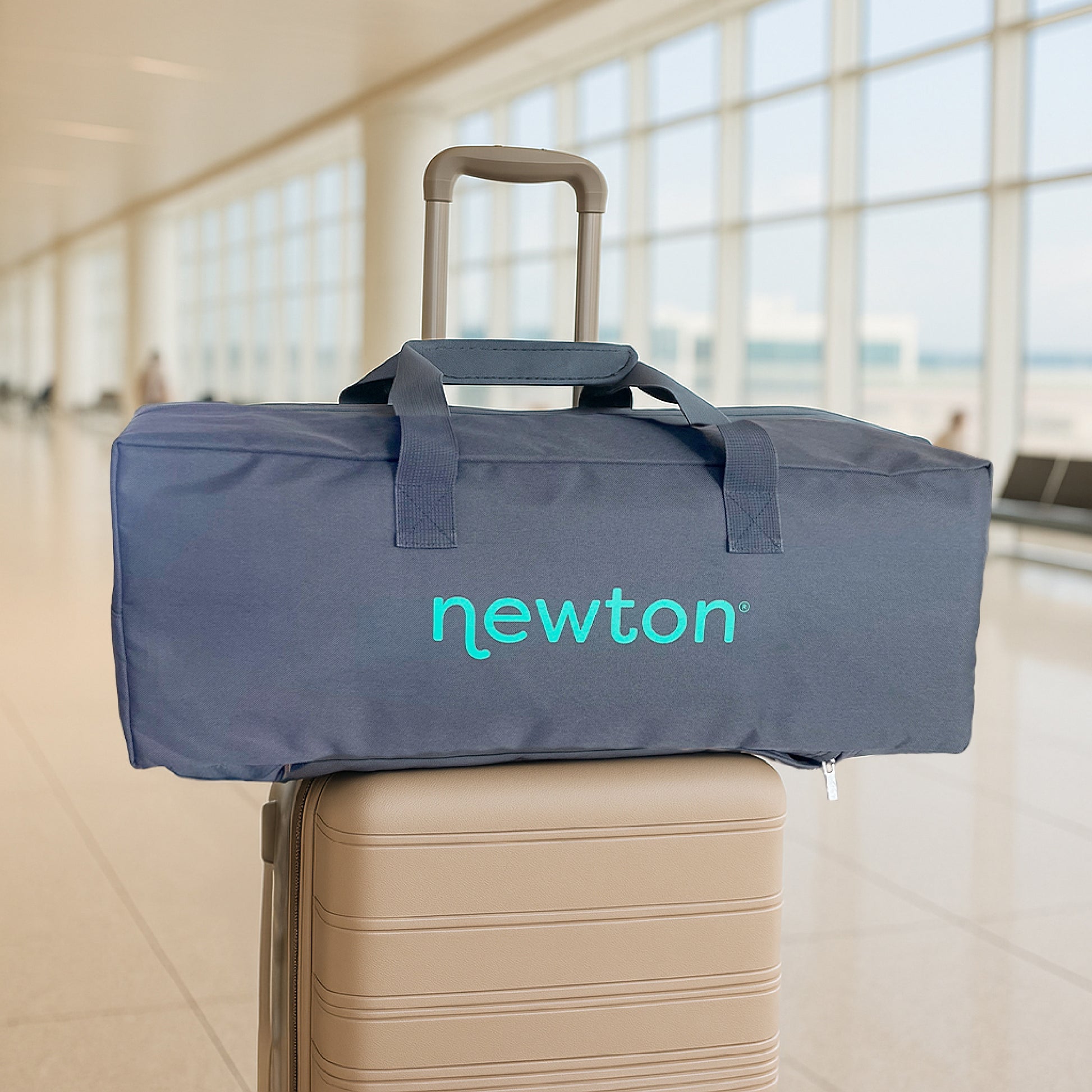 A gray duffel with "Newton Baby" in teal sits on a beige suitcase in a modern airport, perfect for carrying your Compact Travel Crib & Play Yard on the go.