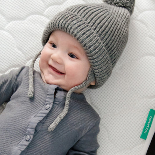 A smiling baby in a grey knit hat and onesie lies on the Newton Baby Waterproof Crib Mattress Pad with a green tag—a breathable, comfy choice for naps or potty training.