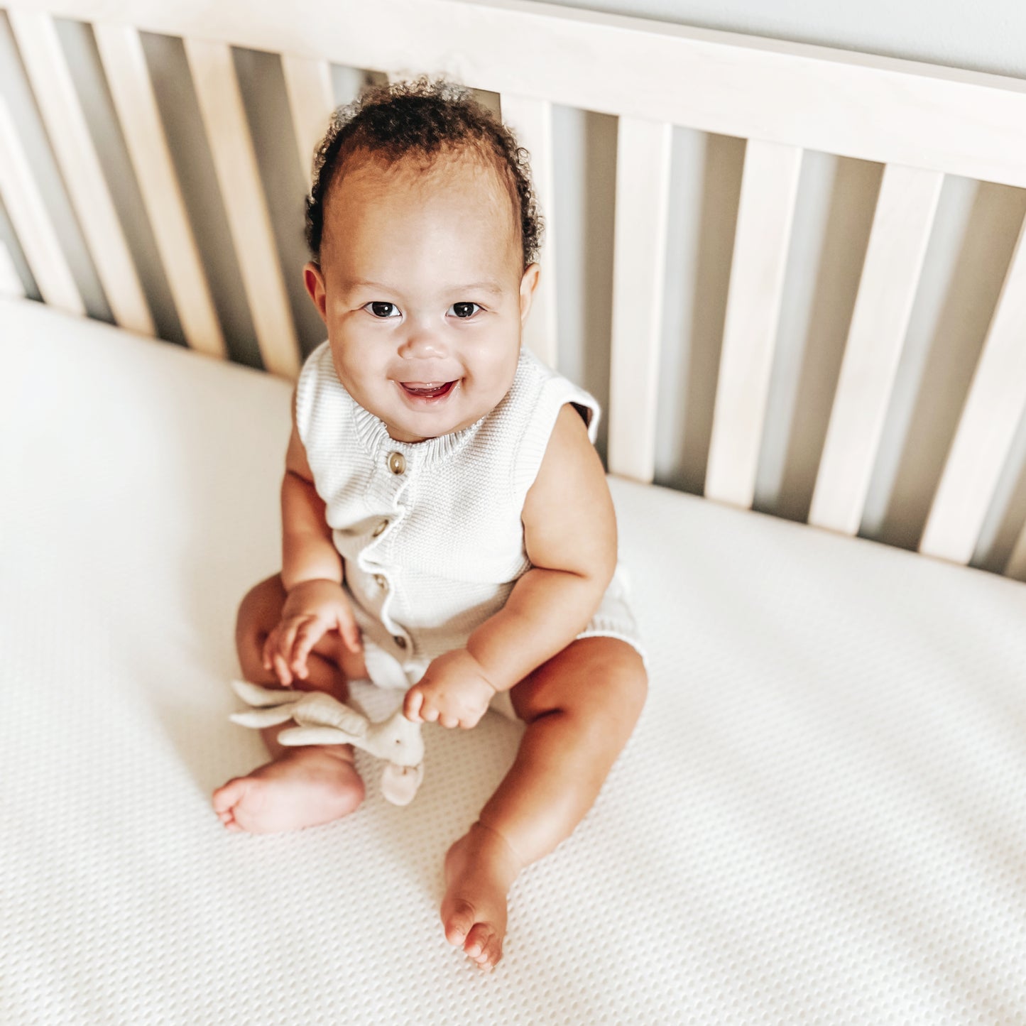 A cheerful baby sitting on a breathable crib mattress, holding a small plush toy.