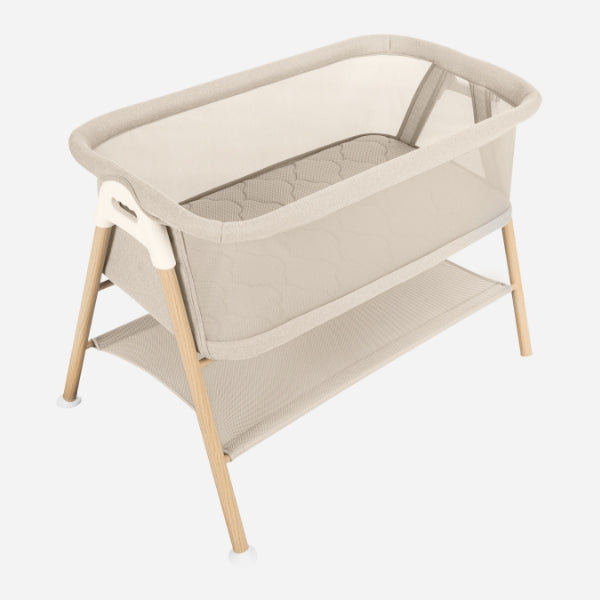 The Newton Baby Essential Bassinet, in beige, features mesh sides, wooden legs, a lower storage shelf, and a breathe-thru mattress. GREENGUARD Gold Certified. Shown against a white background.