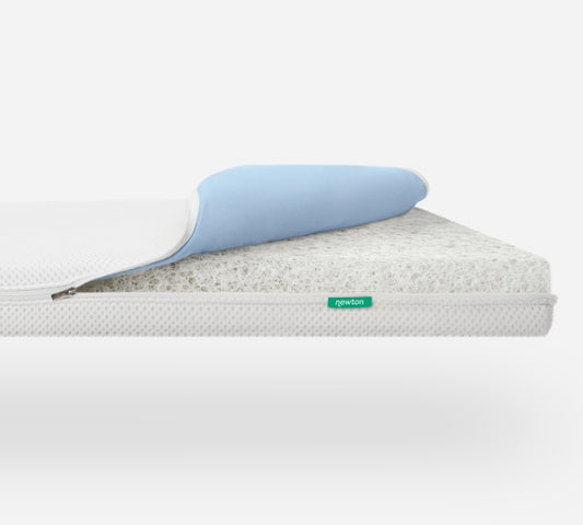 The Newton Baby Essential Waterproof Crib Mattress is shown with its zip-off white cover partially removed, revealing the inner core and blue waterproof mattress pad, all displayed on a plain white background.