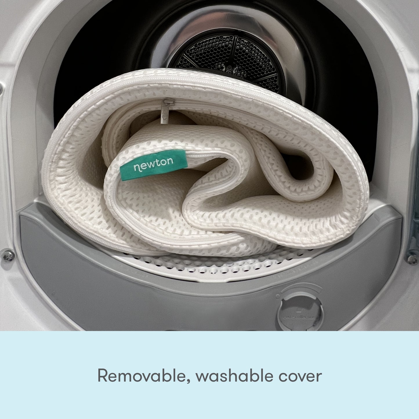 A white folded fabric cover with a green Newton Baby tag is inside an open washer. Text below: Removable, washable cover for the Extra Essential Crib Mattress by Newton Baby.