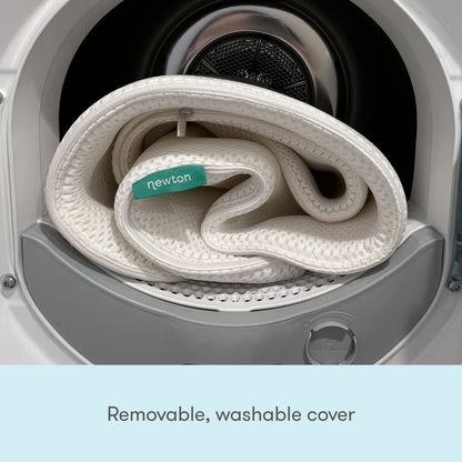 A white folded fabric cover with a green Newton Baby tag is inside an open washer. Text below: Removable, washable cover for the Extra Essential Crib Mattress by Newton Baby.