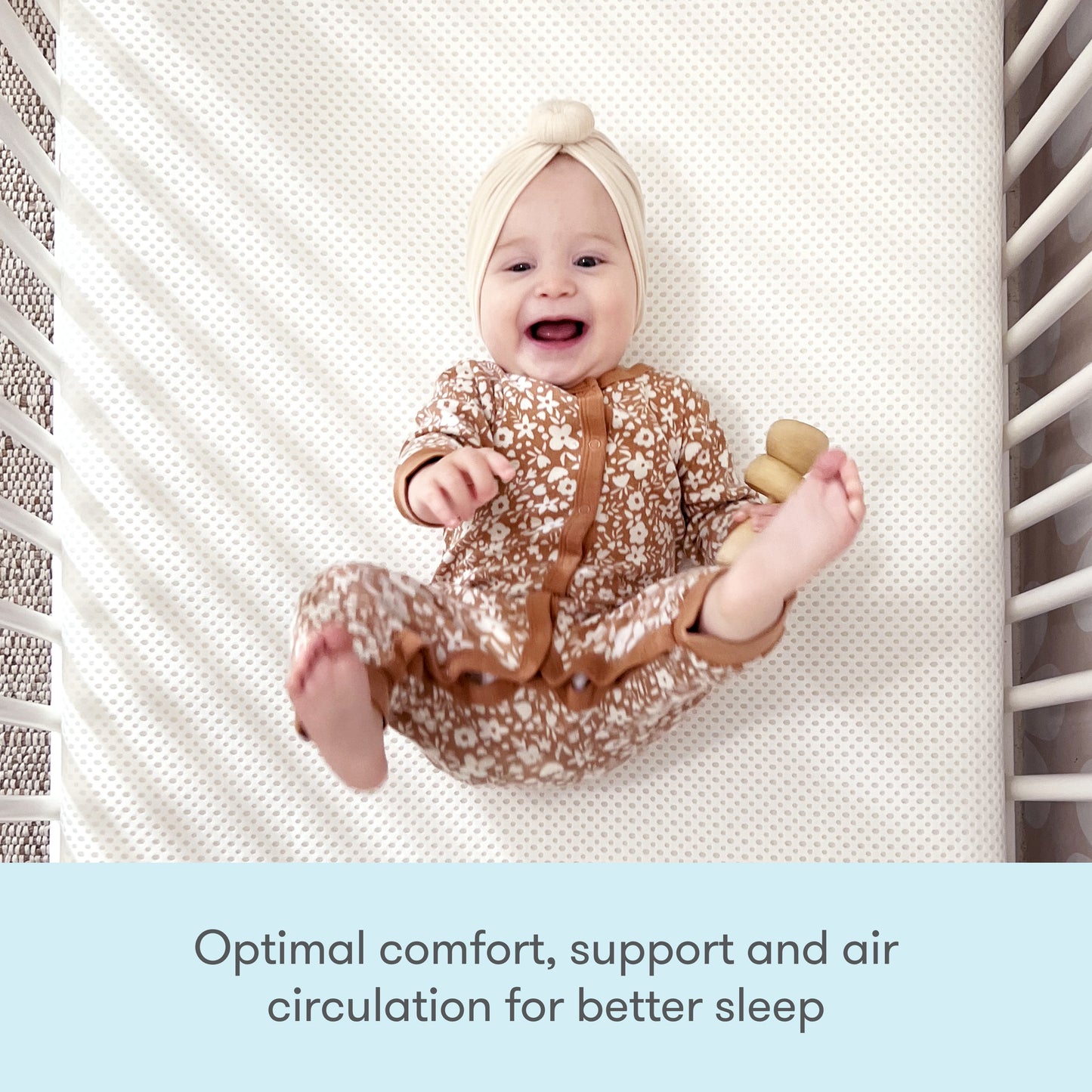 A smiling baby in a brown floral onesie and cream headband lies on a patterned Extra Essential Crib Mattress Cover by Newton Baby, holding a wooden toy. Text below reads, Washable, removable, and designed for optimal comfort and support.