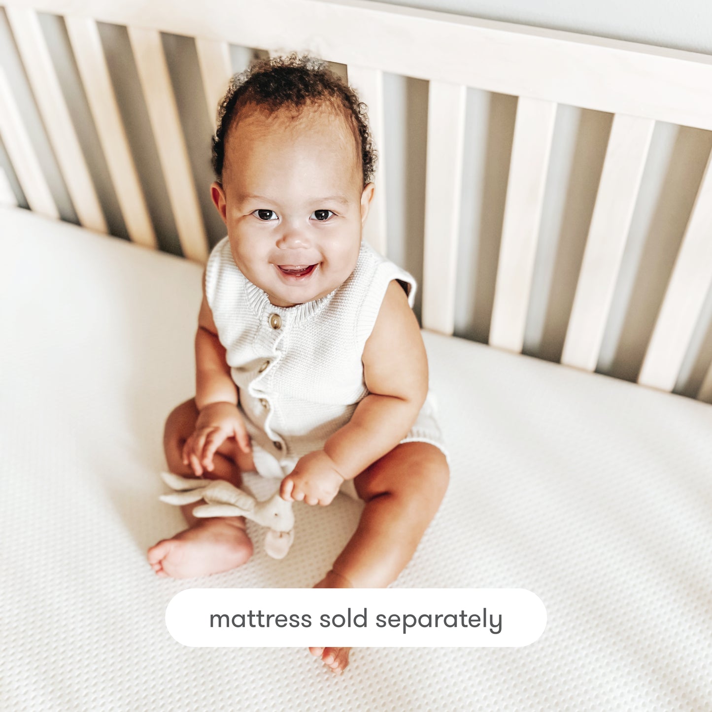 A smiling baby sits on a crib mattress, holding a soft toy inside a light wooden crib. Text at the bottom reads: Newton Baby Extra Essential Crib Mattress Cover, washable and removable, sold separately.