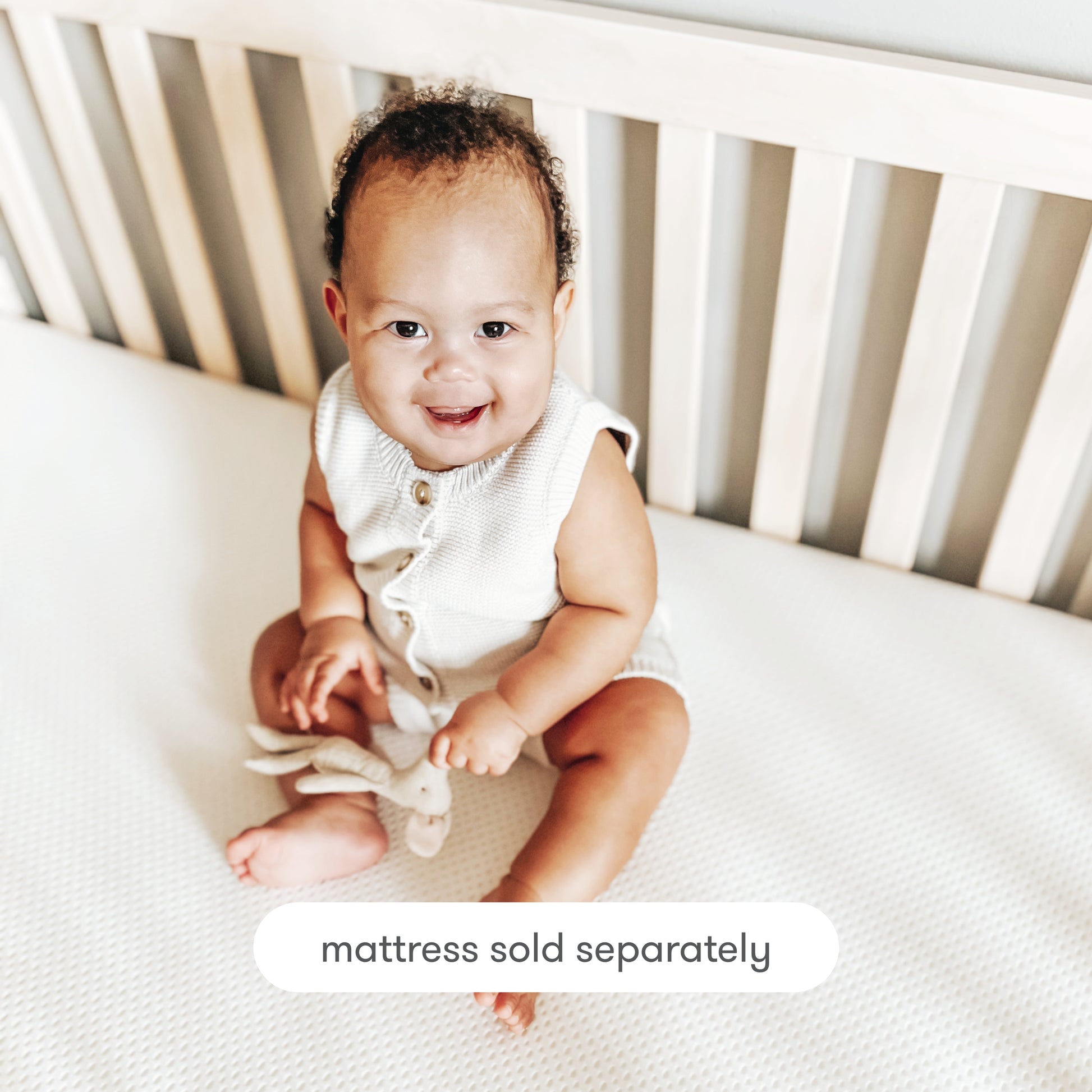 A smiling baby sits on a crib mattress, holding a soft toy inside a light wooden crib. Text at the bottom reads: Newton Baby Extra Essential Crib Mattress Cover, washable and removable, sold separately.