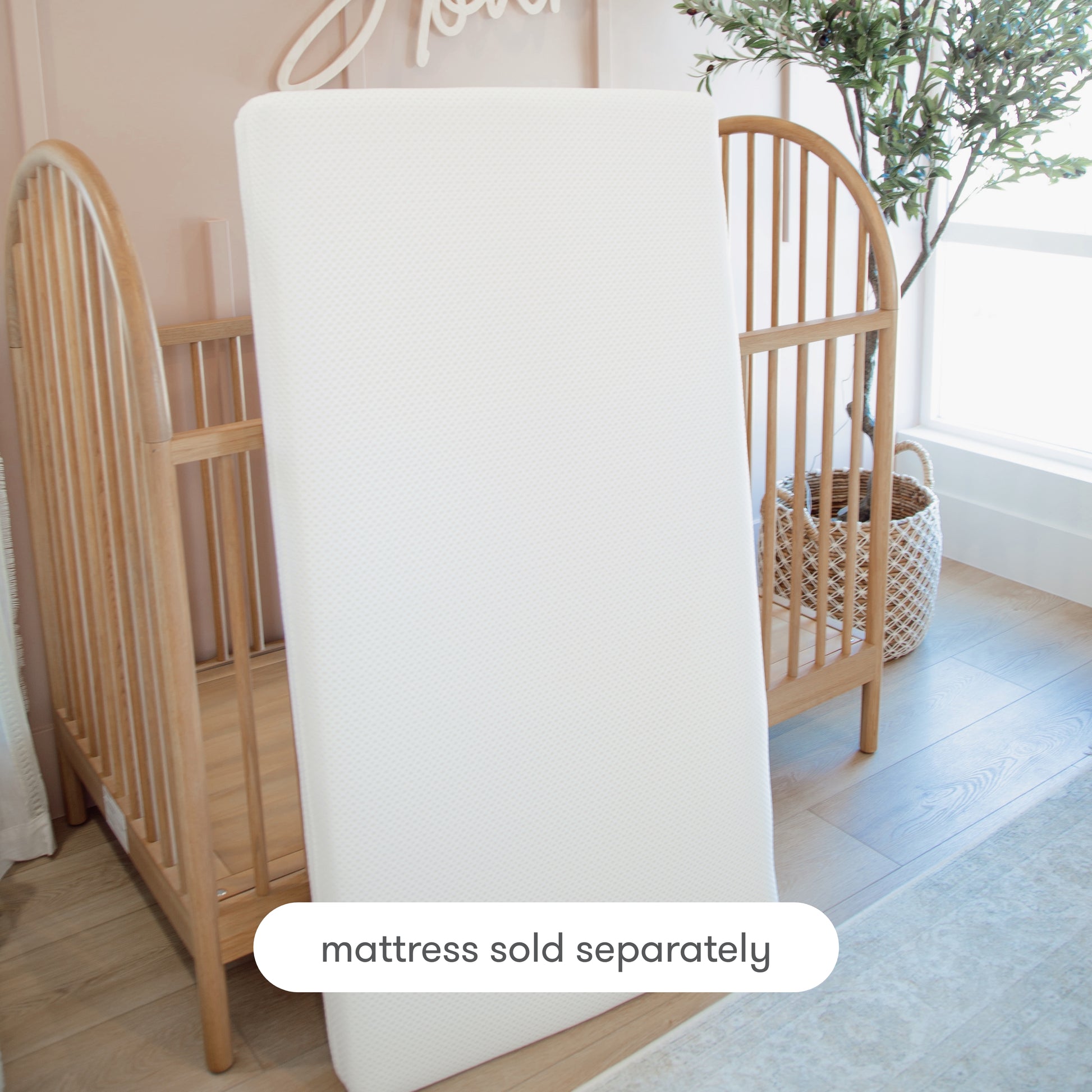 A Newton Baby Extra Essential Crib Mattress Cover is shown upright before a wooden crib in a softly lit nursery. Text at the bottom reads “mattress sold separately.” A plant and woven basket are in the background.