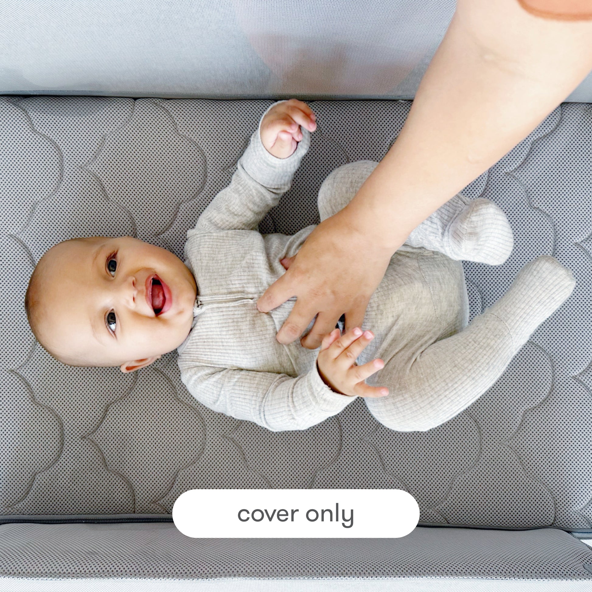 A smiling baby in a gray onesie lies on a quilted Newton Baby Extra Bassinet Mattress Cover while an adult hand gently touches the baby's belly. A label reads "cover only," showing this mattress cover is machine washable.