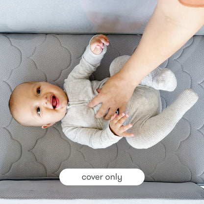 A smiling baby in a gray onesie lies on a quilted Newton Baby Extra Bassinet Mattress Cover while an adult hand gently touches the baby's belly. A label reads "cover only," showing this mattress cover is machine washable.
