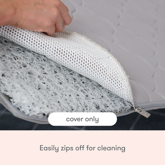 A hand unzips the textured, light-colored Extra Bassinet Mattress Cover from Newton Baby, revealing the inside. Text on image reads: "Bassinet mattress cover only. Easily zips off for machine washable cleaning.