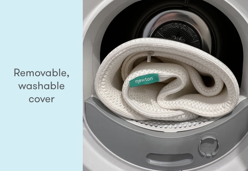 Newton baby mattress cover coiled inside a washing machine with the label visible, and text stating "Removable, washable cover."