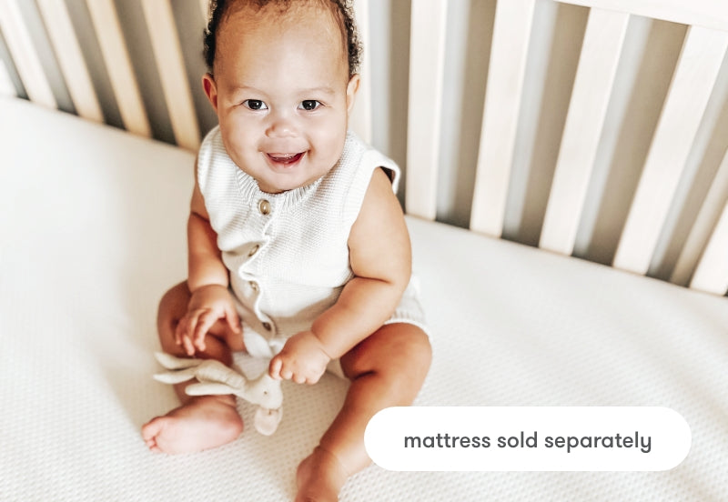 Happy baby sitting on a crib mattress with a toy, text overlay states "mattress sold separately".