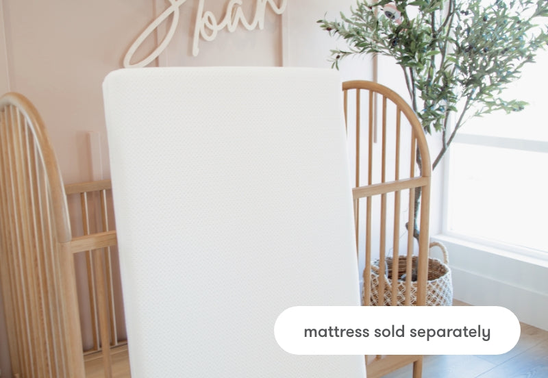 Crib mattress displayed vertically in a nursery room with a soft color scheme, accompanied by a label indicating "mattress sold separately."