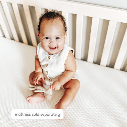 A smiling baby in a white romper sits in a crib with light wooden slats, on a Newton Baby Extra Essential Crib Mattress Cover that's ultra-breathable and washable. Text on the image reads: mattress sold separately.
