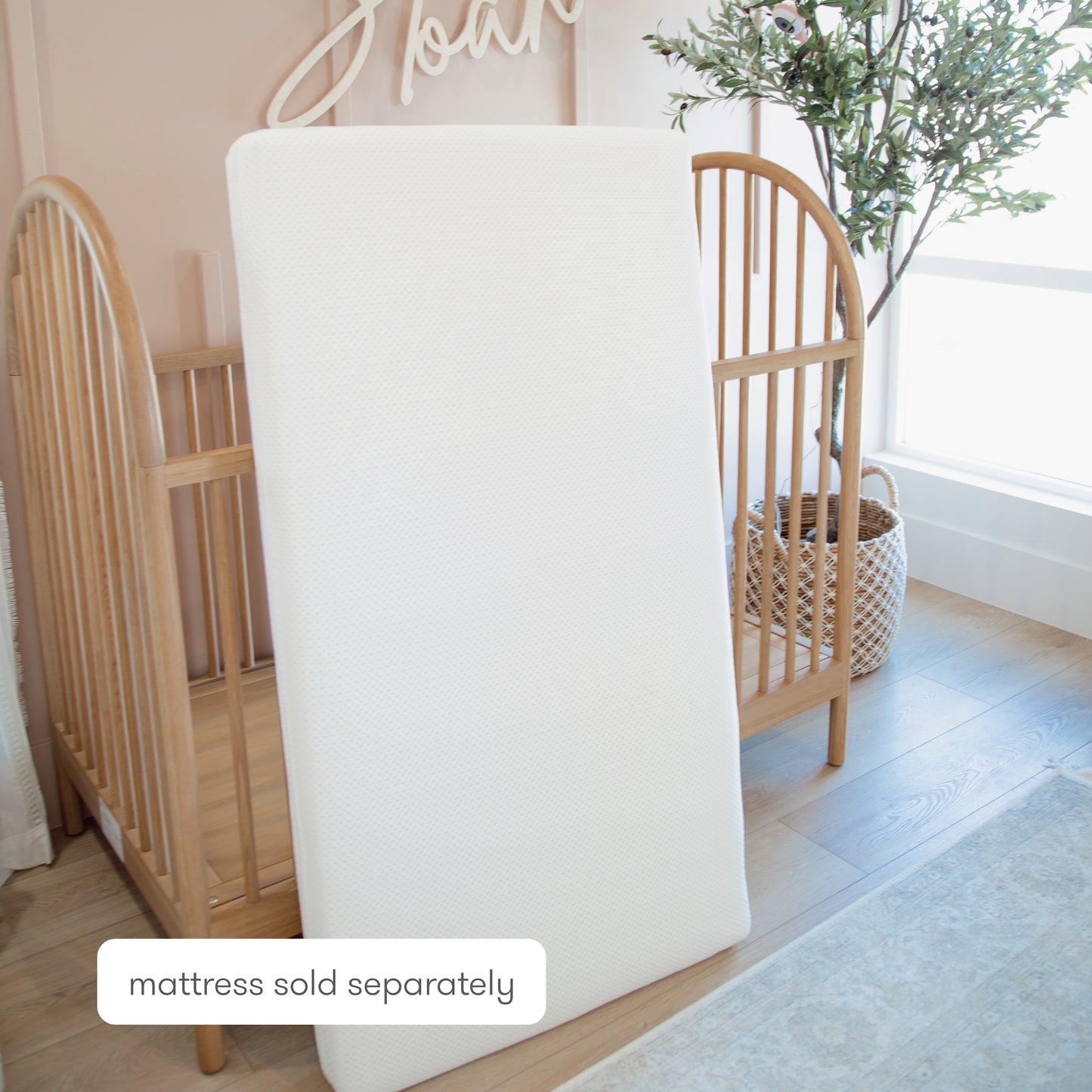 A white, ultra-breathable Newton Baby Extra Essential Crib Mattress Cover stands upright before a wooden crib in a softly lit nursery. A plant in a woven basket is by the window. Text reads: mattress sold separately.