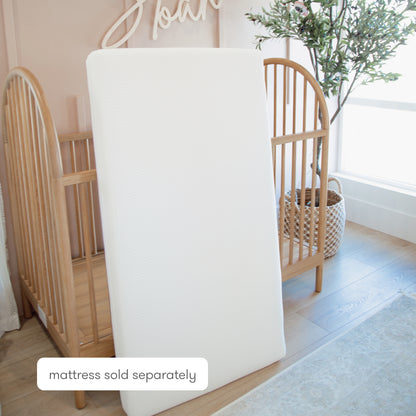 A white, ultra-breathable Newton Baby Extra Essential Crib Mattress Cover stands upright before a wooden crib in a softly lit nursery. A plant in a woven basket is by the window. Text reads: mattress sold separately.