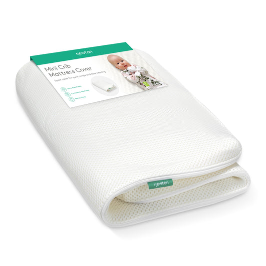 A folded white Extra Mini Crib Mattress Cover by Newton Baby with a green and white label featuring a baby and product details. This washable cover is designed for comfort and convenience.