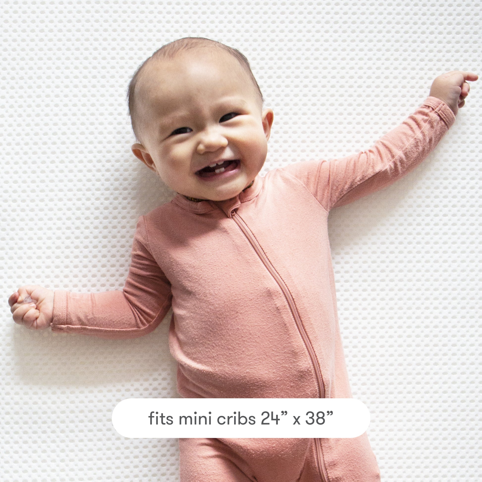 A smiling baby in pink lies on the Newton Baby Extra Mini Crib Mattress Cover, a white, breathable, textured protector made to fit mini cribs sized 24" x 38".