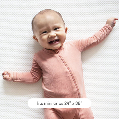 A smiling baby in pink lies on the Newton Baby Extra Mini Crib Mattress Cover, a white, breathable, textured protector made to fit mini cribs sized 24" x 38".