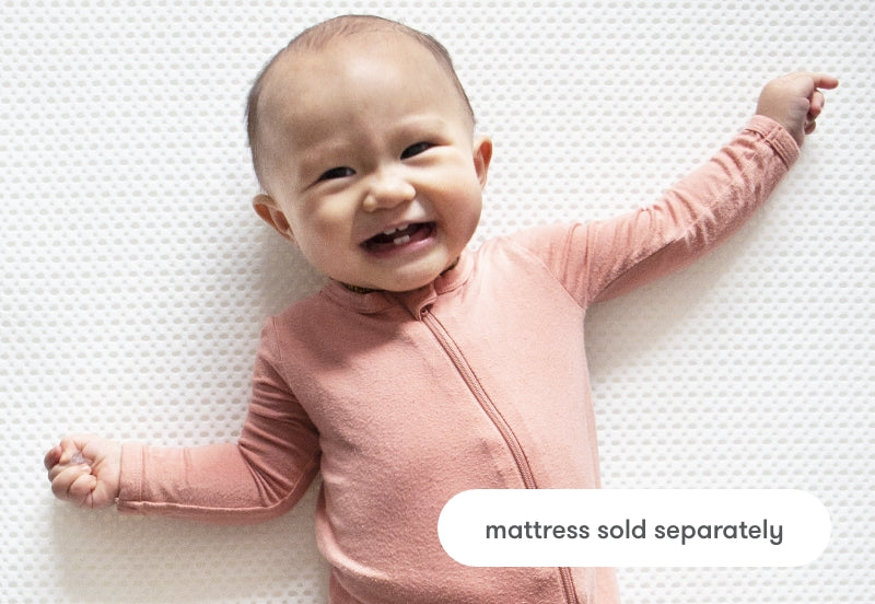 A smiling baby in a pink onesie lying on a textured white surface with the text "mattress sold separately."
