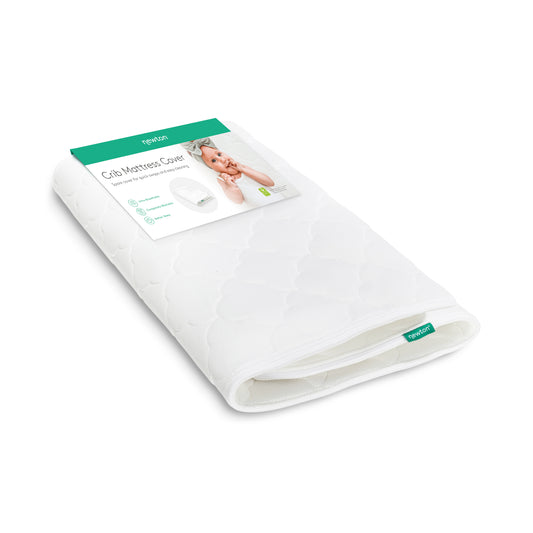 A washable white Extra Original Crib Mattress Cover with a quilted pattern is shown folded. The packaging displays a baby photo and product details, with the Newton Baby brand name clearly visible on the label and tag.