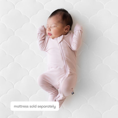 A newborn baby in a light pink onesie sleeps on the Newton Baby Extra Original Crib Mattress Cover, which is ultra-breathable with a white, patterned design. Note: Mattress sold separately.