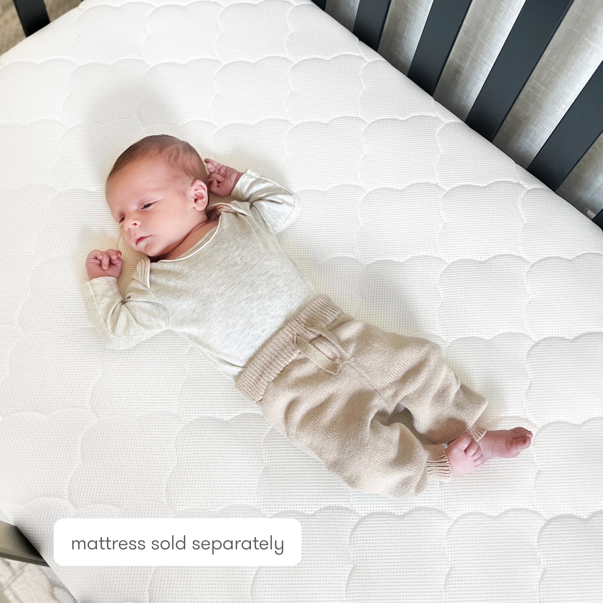 A newborn in a beige outfit rests on a Newton Baby Extra Original Crib Mattress Cover with a soft cloud-like pattern. The ultra-breathable, washable cover ensures comfort. Text on image: mattress sold separately.