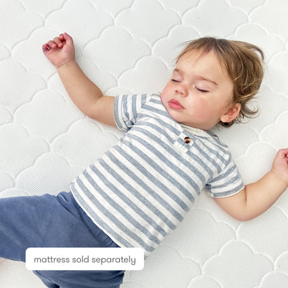 A toddler in a gray and white striped shirt and blue pants sleeps on the Newton Baby Extra Original Crib Mattress Cover, which is ultra-breathable and white. A label at the bottom states: mattress sold separately.