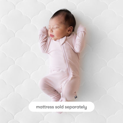 A newborn baby in a light pink zip-up onesie sleeps on a white crib mattress featuring the Newton Baby Extra Original Crib Mattress Cover. Text at the bottom reads, “mattress sold separately.”.
