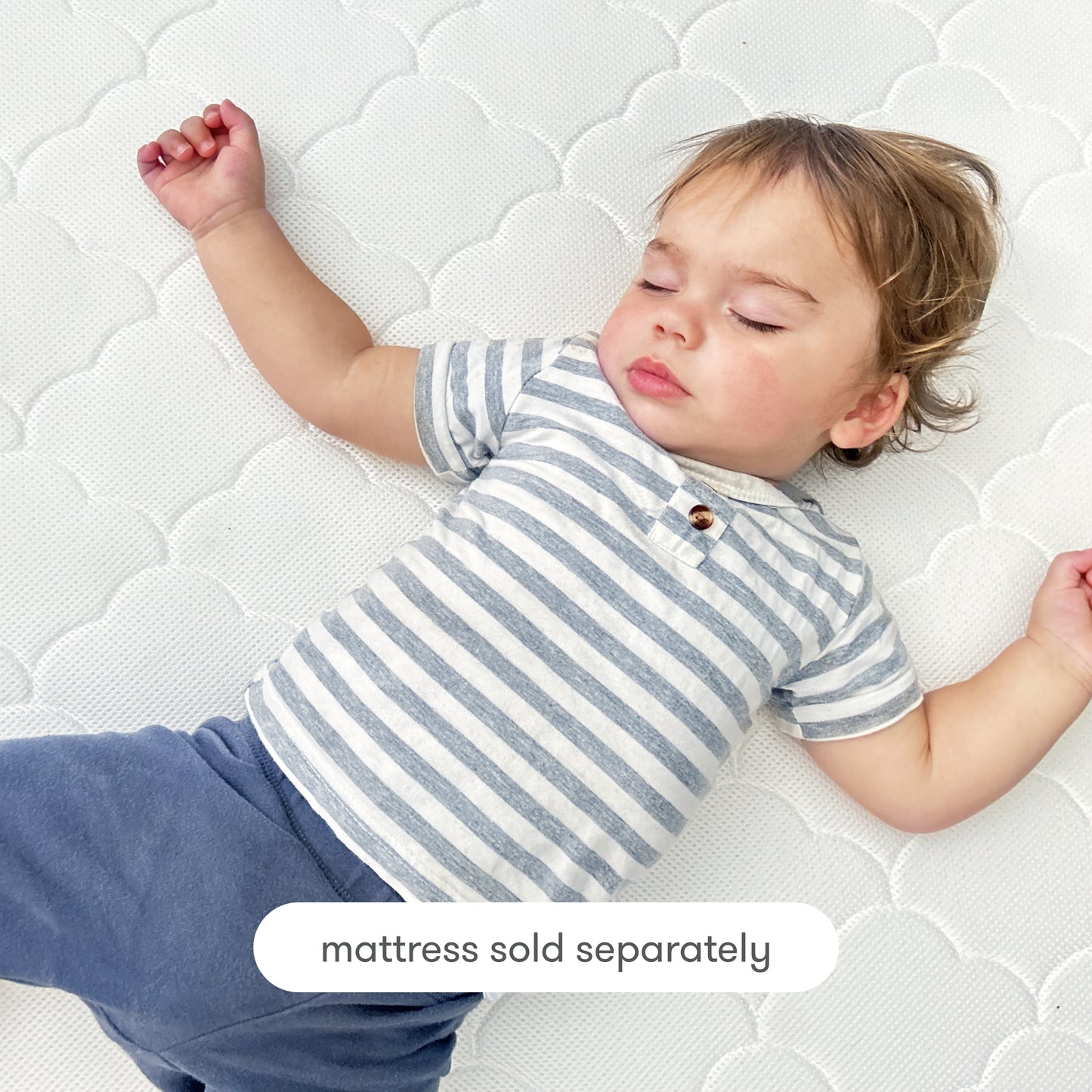 A toddler with light brown hair, wearing a gray and white striped shirt and blue pants, sleeps on a white patterned breathable Newton Baby Extra Original Crib Mattress Cover. Text at the bottom reads, mattress sold separately.