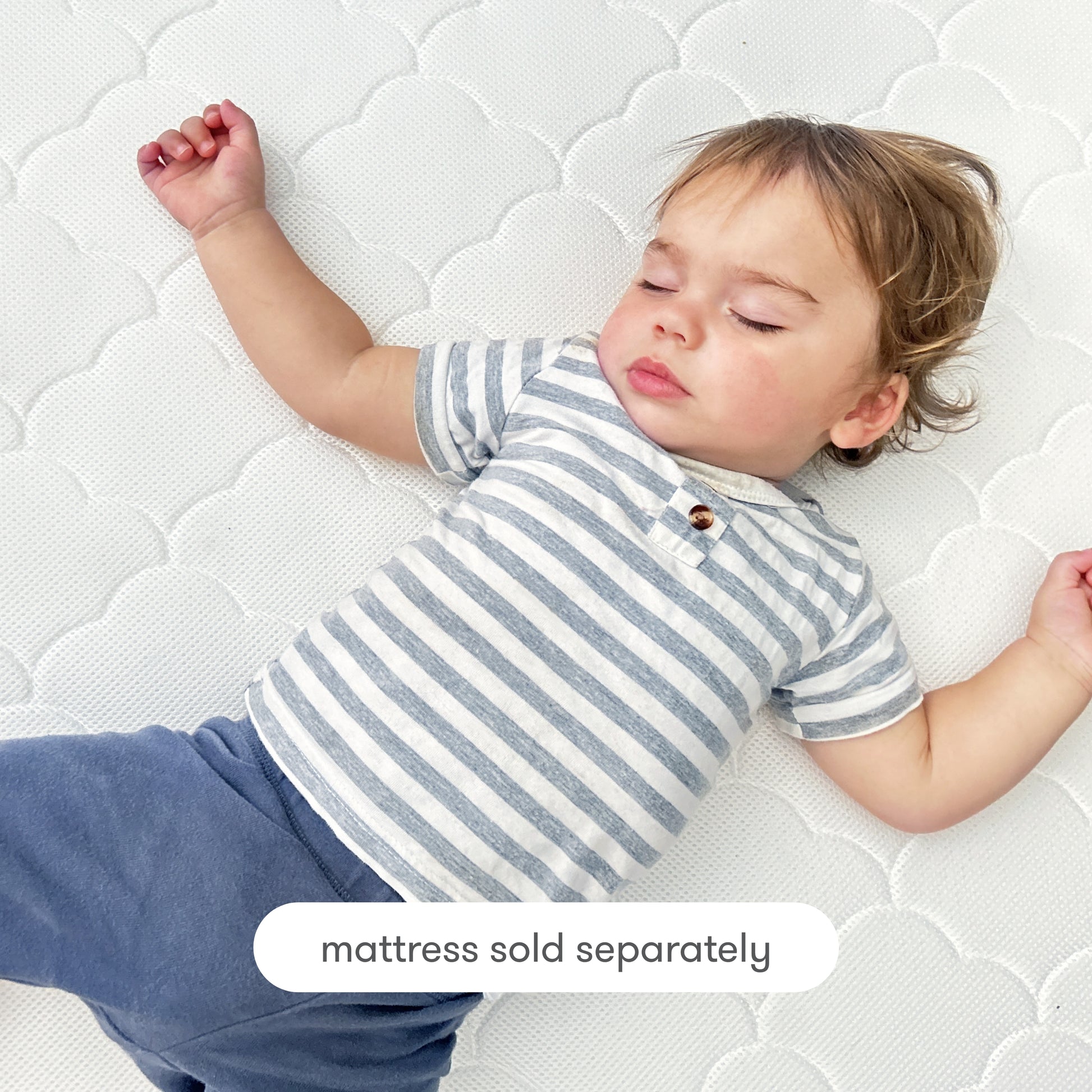 A toddler with light brown hair, wearing a gray and white striped shirt and blue pants, sleeps on a white patterned breathable Newton Baby Extra Original Crib Mattress Cover. Text at the bottom reads, mattress sold separately.