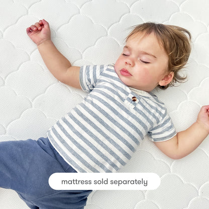 A toddler with light brown hair, wearing a gray and white striped shirt and blue pants, sleeps on a white patterned breathable Newton Baby Extra Original Crib Mattress Cover. Text at the bottom reads, mattress sold separately.