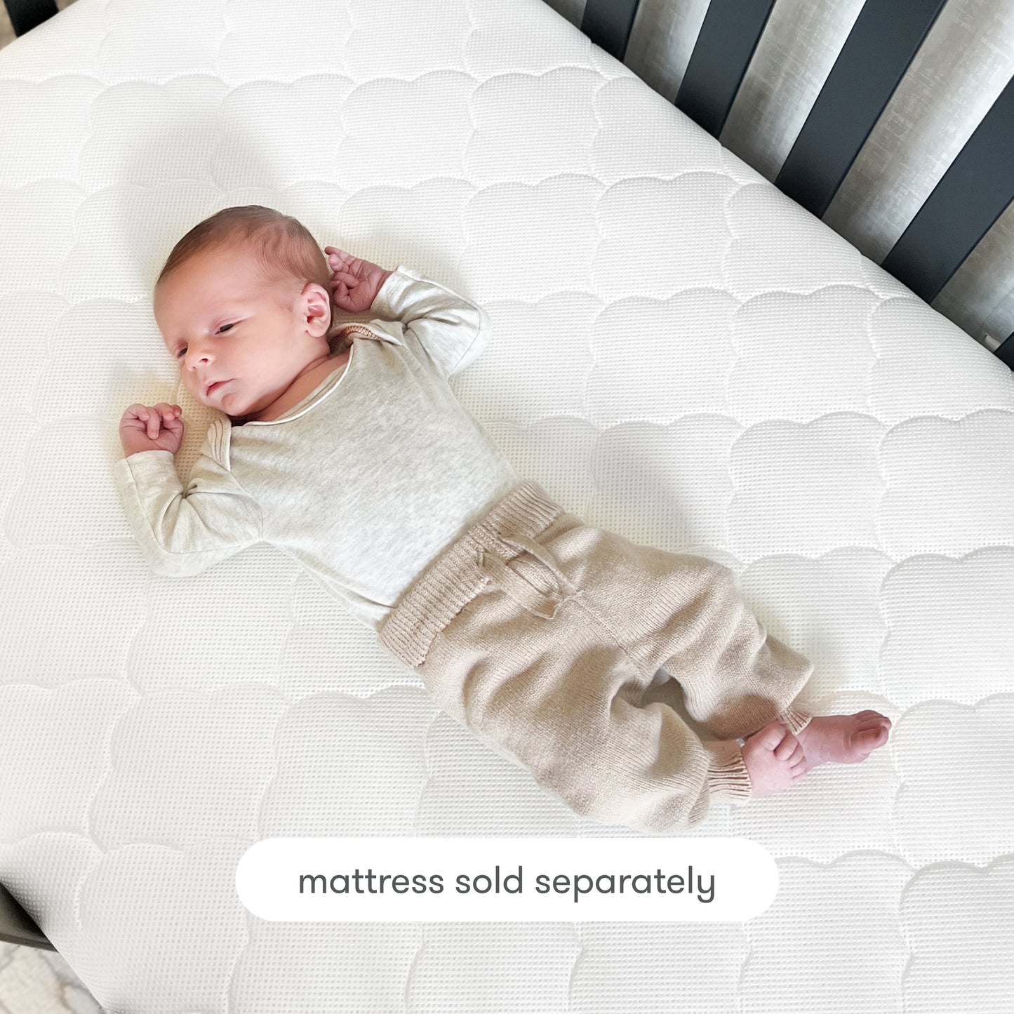 A baby in a cream long-sleeve shirt and beige pants lies on a white crib mattress topped with the Newton Baby Extra Original Crib Mattress Cover. Text at the bottom reads, mattress sold separately.