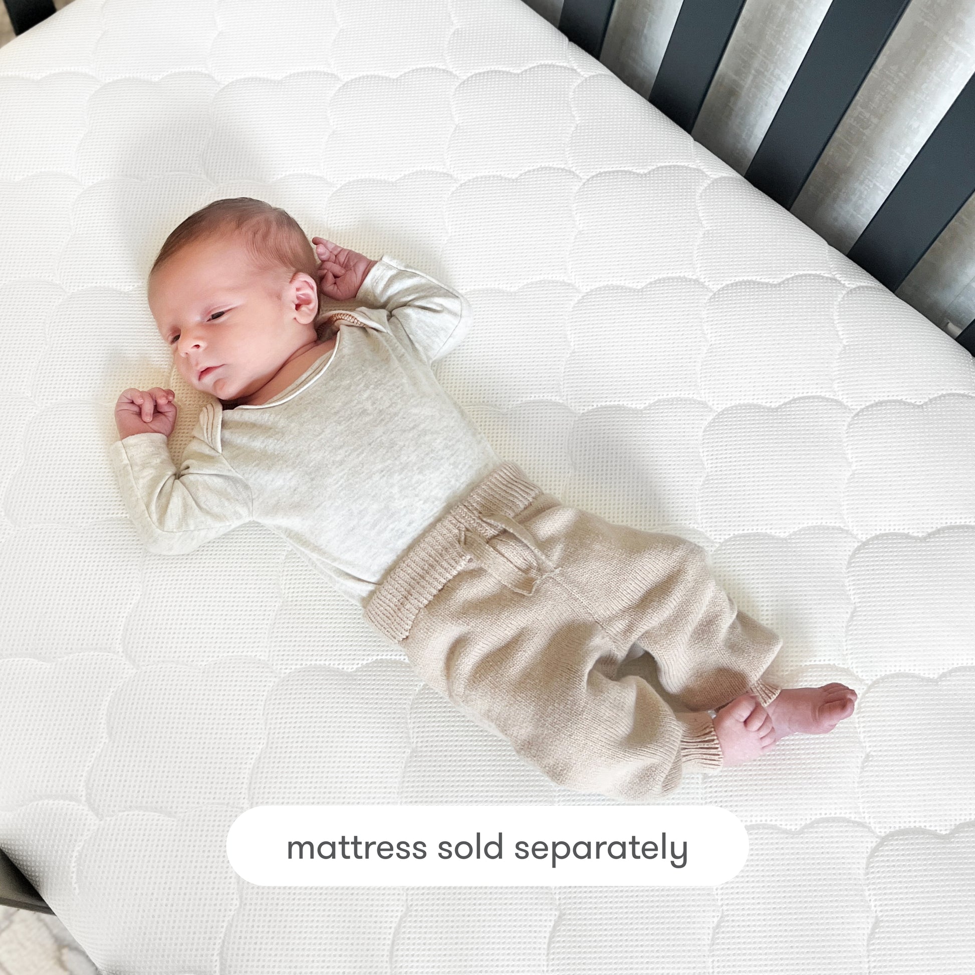 A baby in a cream long-sleeve shirt and beige pants lies on a white crib mattress topped with the Newton Baby Extra Original Crib Mattress Cover. Text at the bottom reads, mattress sold separately.