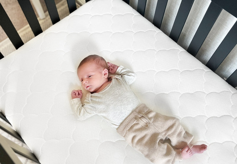 A peaceful newborn lies comfortably on a white patterned mattress inside a dark crib, wearing a cozy beige outfit.