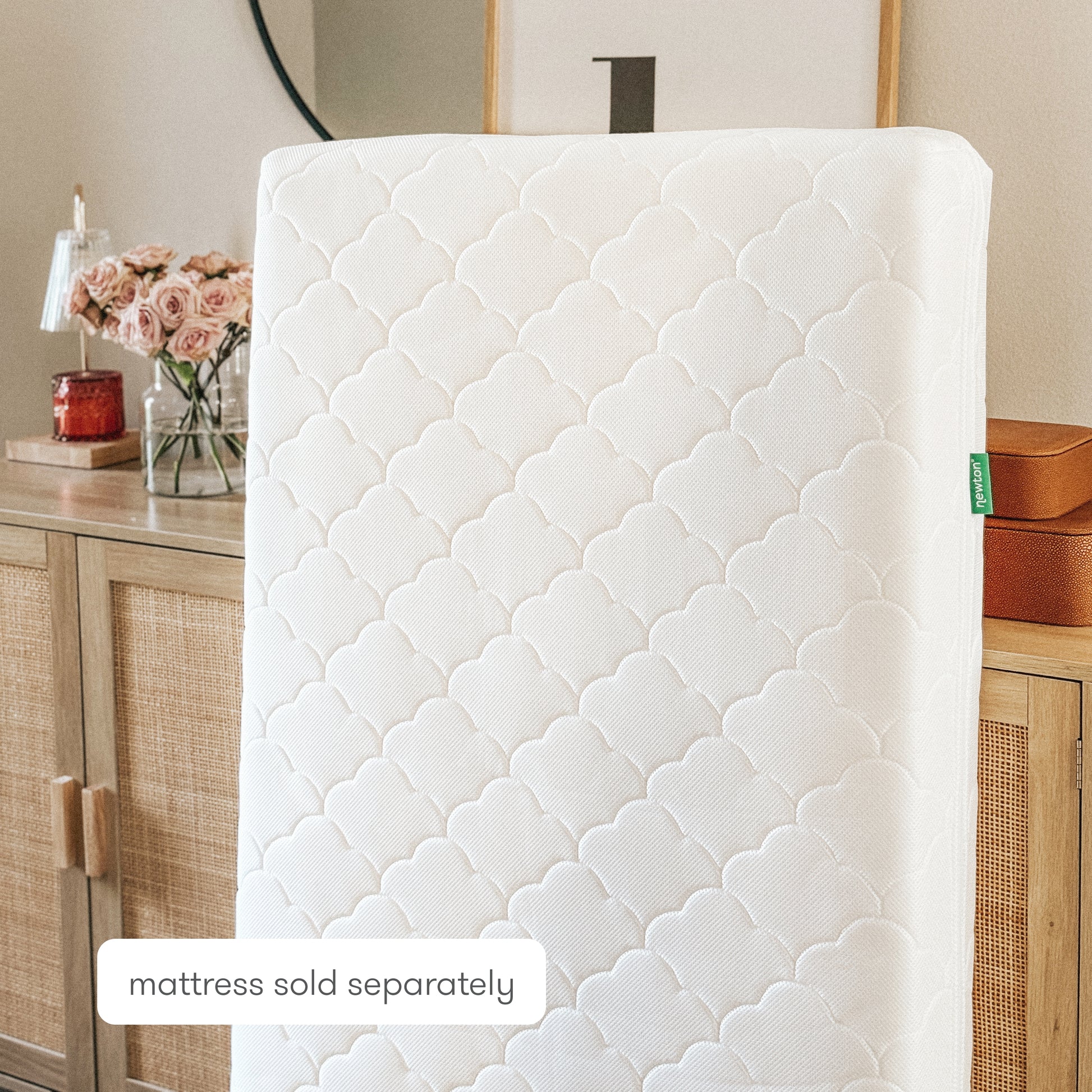 A white, quilted Newton Baby Extra Waterproof Crib Mattress Cover stands upright in front of a wooden dresser with rattan doors. A vase of pink roses and home decor are visible. Text reads mattress sold separately.