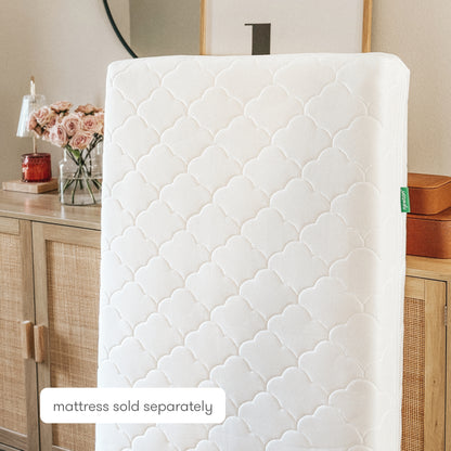 A white, quilted Newton Baby Extra Waterproof Crib Mattress Cover stands upright in front of a wooden dresser with rattan doors. A vase of pink roses and home decor are visible. Text reads mattress sold separately.